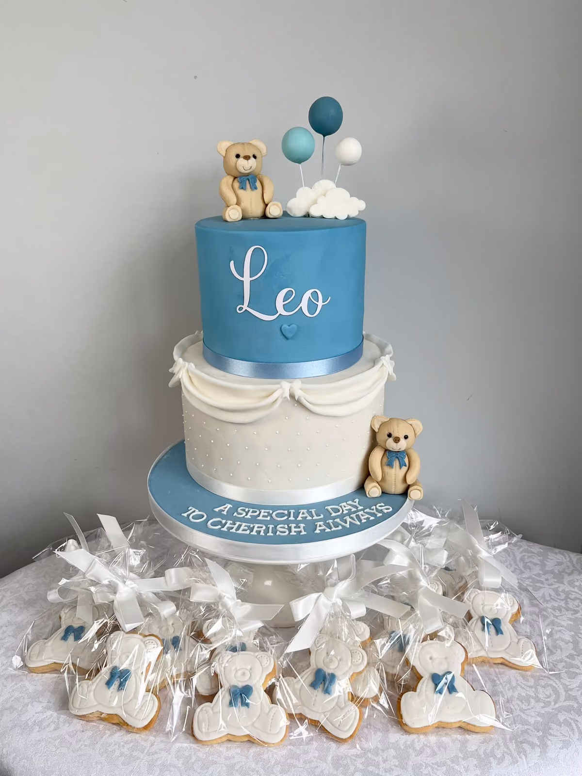 A two-tier blue and white christening cake for Leo with sugar teddy bears, blue balloons and a cloud, reading 'A special day to cherish always', surrounded by wrapped teddy bear biscuits.