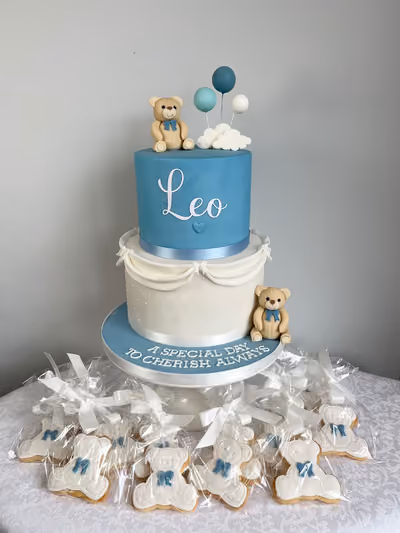 A two-tier blue and white christening cake for Leo with sugar teddy bears, blue balloons and a cloud, reading 'A special day to cherish always', surrounded by wrapped teddy bear biscuits.