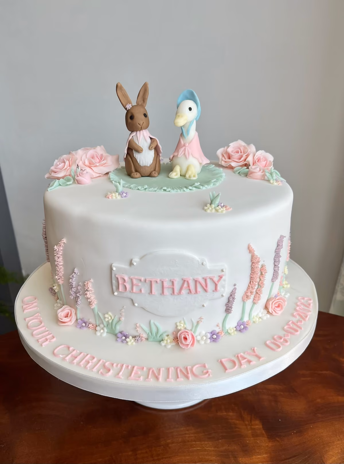 A white christening cake decorated with fondant figures of a brown rabbit and a white duck, surrounded by pink roses and various sugar flowers. The cake has 'BETHANY' on a plaque on the side and 'ON YOUR CHRISTENING DAY 09-07-2023' written in pink letters around the base.