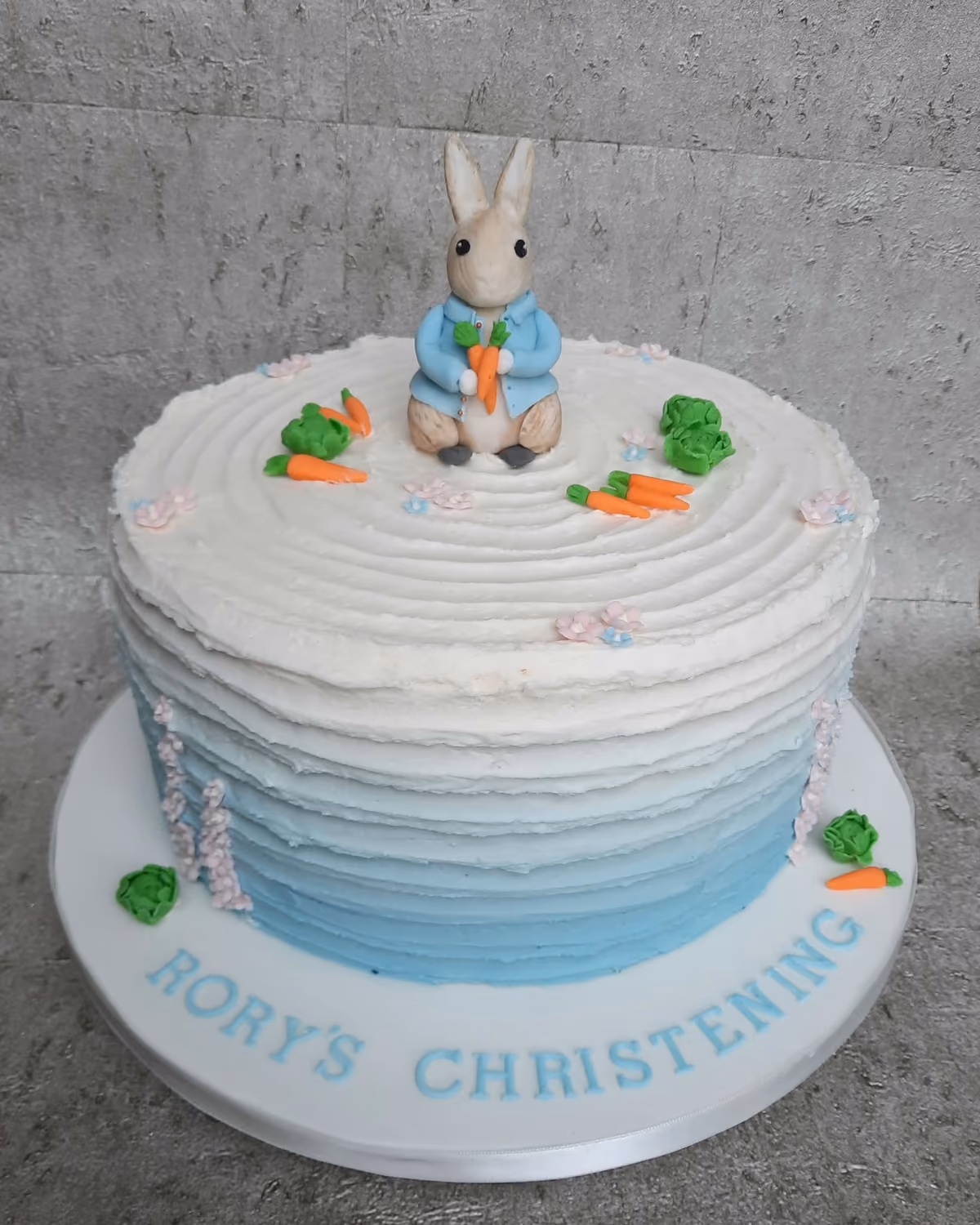 A round christening cake featuring a Peter Rabbit figure holding a carrot on top, with an ombre blue and white frosted side, and small fondant carrots, lettuce, and pink and blue flowers scattered around. The white cake board has 'RORY'S CHRISTENING' written in light blue letters.