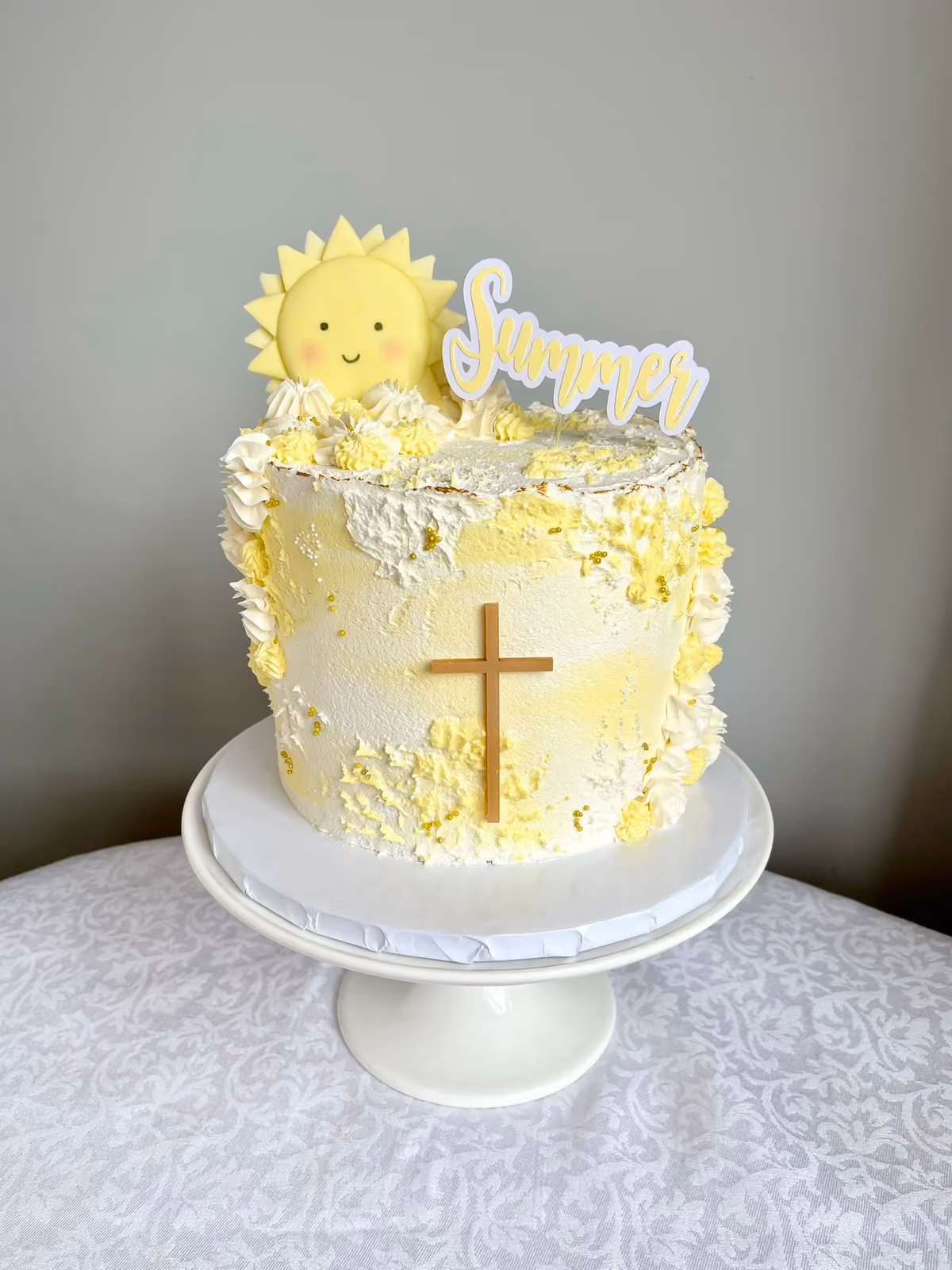 A round, single-tier cake with white and yellow textured frosting, decorated with a smiling yellow sun topper, a 'Summer' cake topper, a small wooden cross, and gold sprinkles, displayed on a white pedestal cake stand.