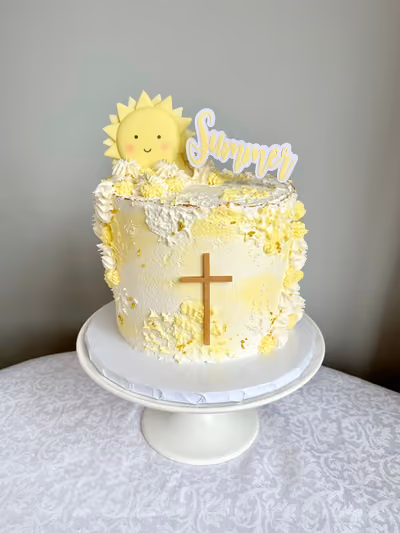 A round, single-tier cake with white and yellow textured frosting, decorated with a smiling yellow sun topper, a 'Summer' cake topper, a small wooden cross, and gold sprinkles, displayed on a white pedestal cake stand.