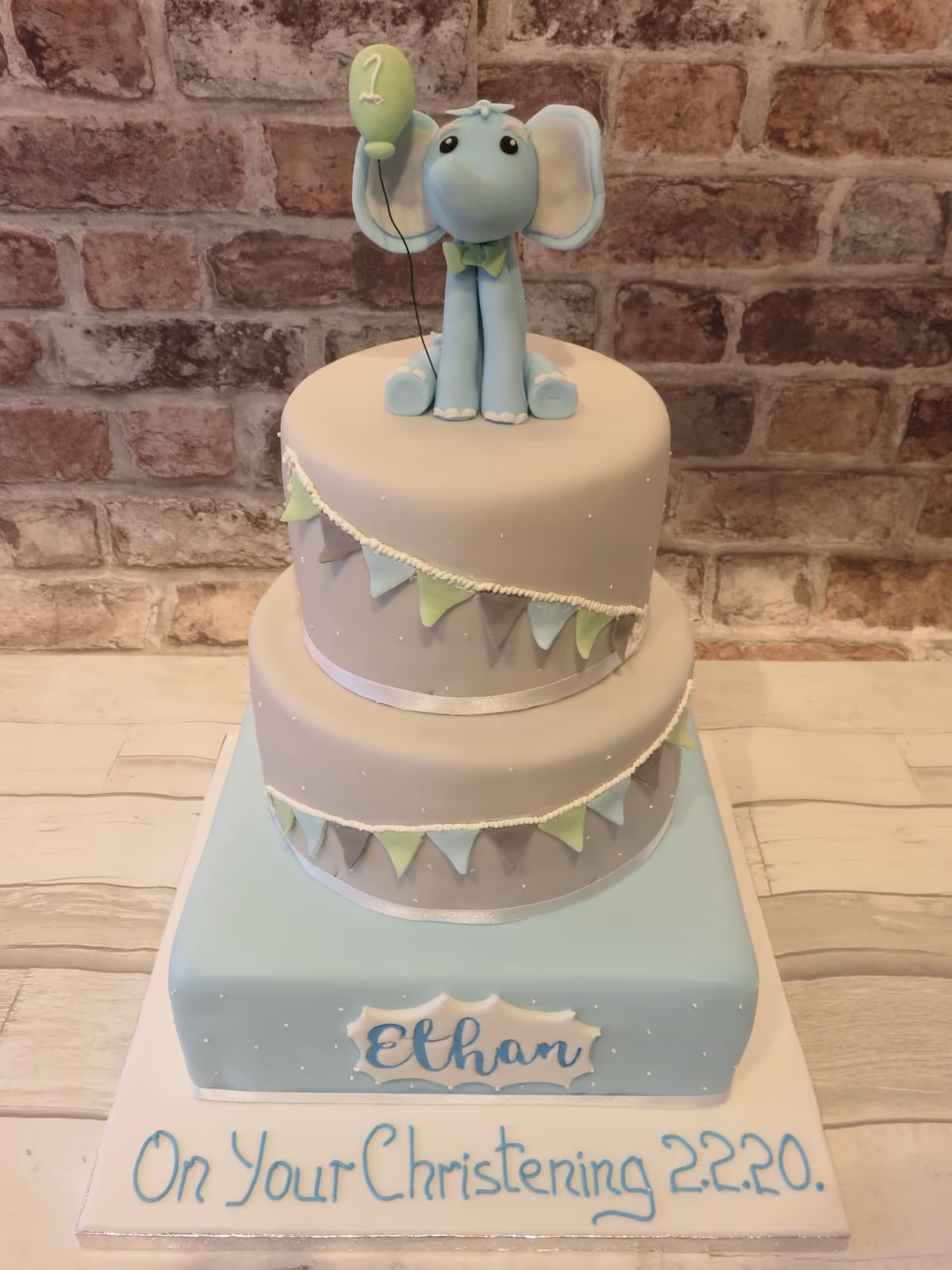 A two-tier christening cake with light grey tiers decorated with pastel bunting and white polka dots, and a light blue base with 'Ethan' written on it. A blue elephant topper holding a green balloon with the number '1' sits on top. The cake board reads 'On Your Christening 2.2.20.'.