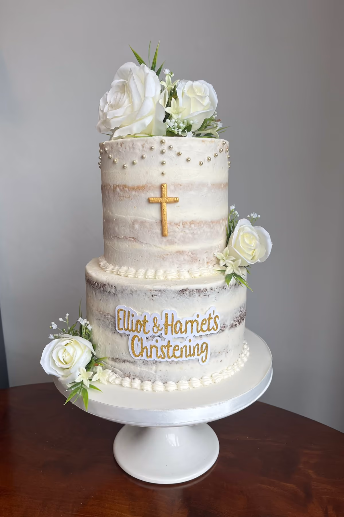 A two-tier naked christening cake decorated with white roses and greenery, golden pearls, a gold cross, and a gold glitter 'Elliot & Harriet's Christening' sign.