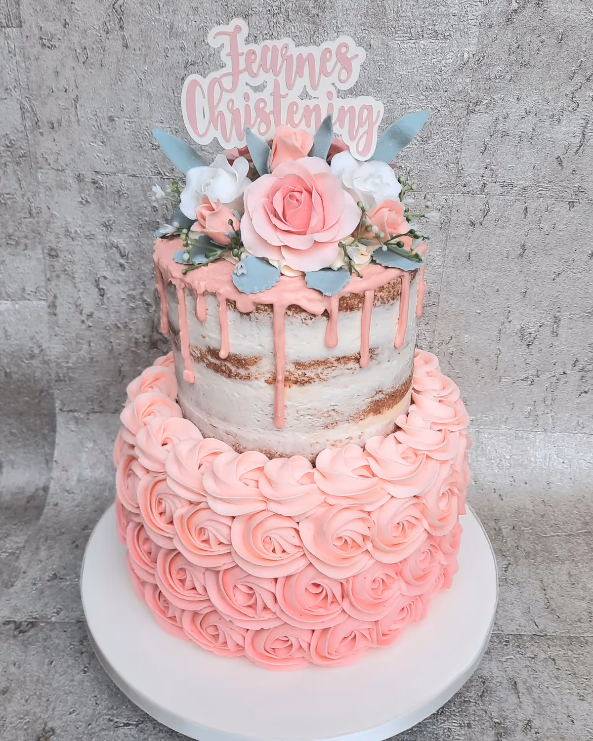 A two-tier christening cake with a 'Feurnes Christening' topper. The top tier is a naked cake with pink drip icing, decorated with pink roses, white flowers, and blue leaves. The bottom tier is covered in pink buttercream rosettes.