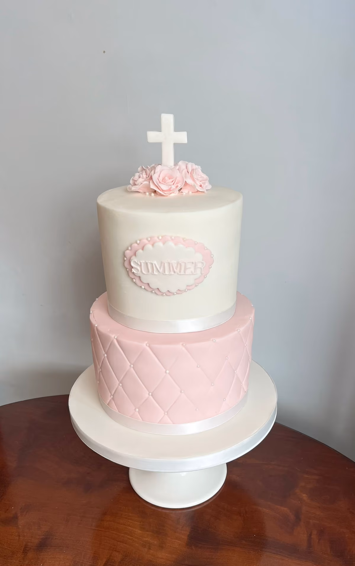 A two-tier christening cake featuring a white top tier with a white cross, pink sugar roses, and a 'SUMMER' name plaque, and a pink bottom tier with a quilted pattern and pearl details, all presented on a white cake stand.