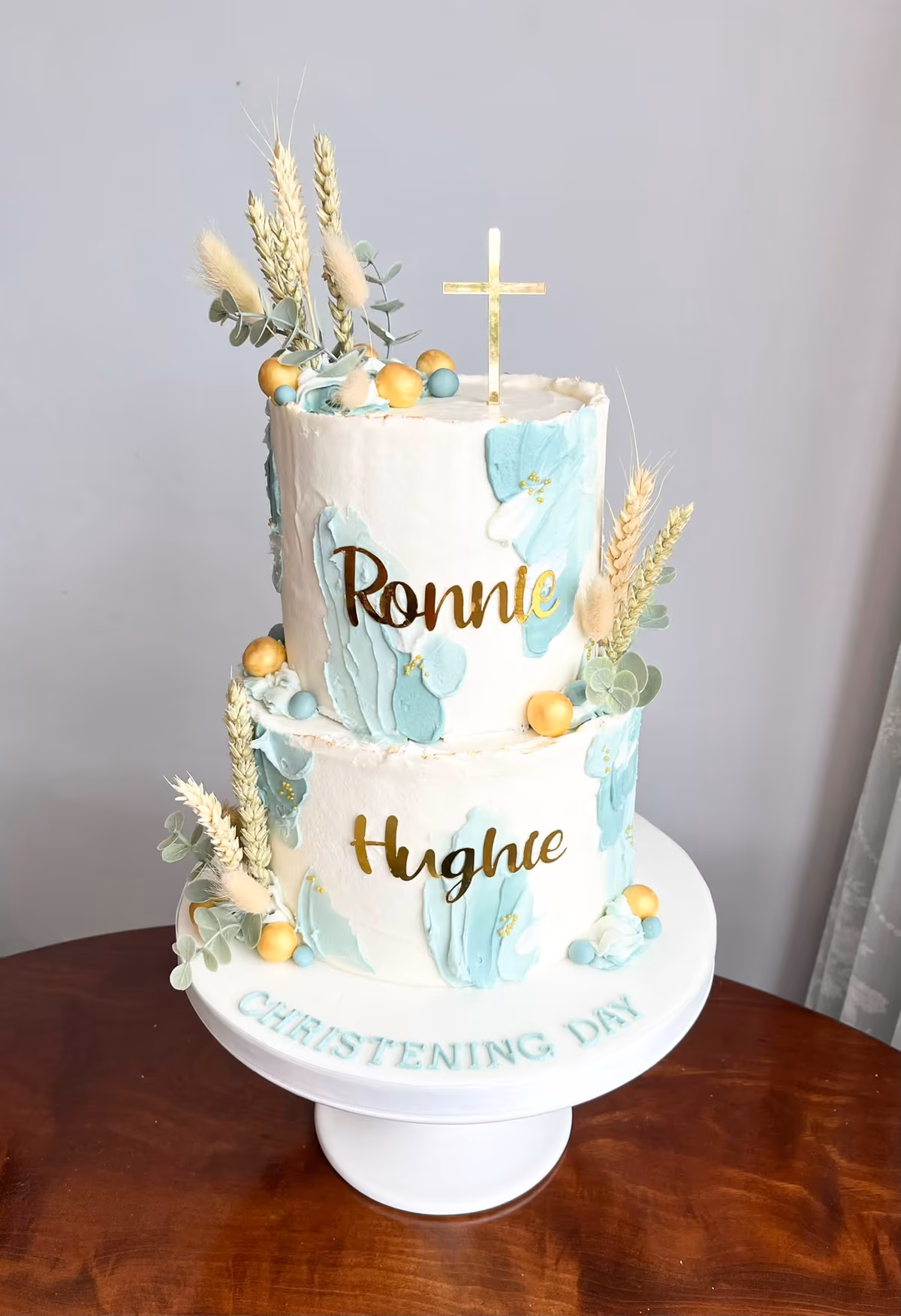 A two-tier white buttercream christening cake with mint green brushstroke accents and gold names 'Ronnie' and 'Hughie'. It is decorated with dried floral arrangements including wheat and eucalyptus, gold spheres, and topped with a gold cross. The cake stand reads 'CHRISTENING DAY' in mint green.