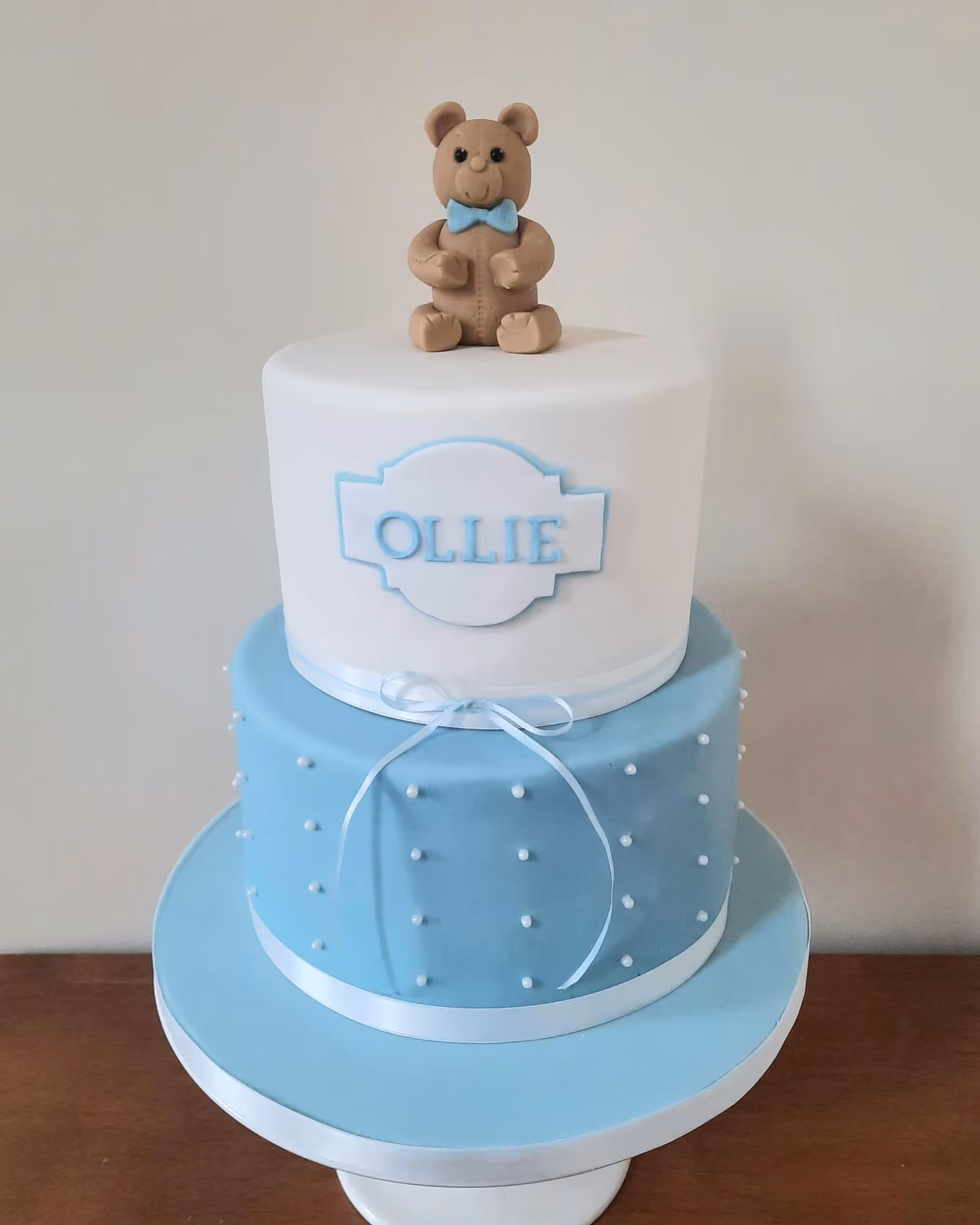 A two-tier christening cake with a white top tier and a light blue bottom tier, adorned with a brown teddy bear topper wearing a blue bow tie. The white tier features a light blue plaque with the name 'OLLIE', and the blue tier is decorated with small white pearl dots and a light blue ribbon tied in a bow.