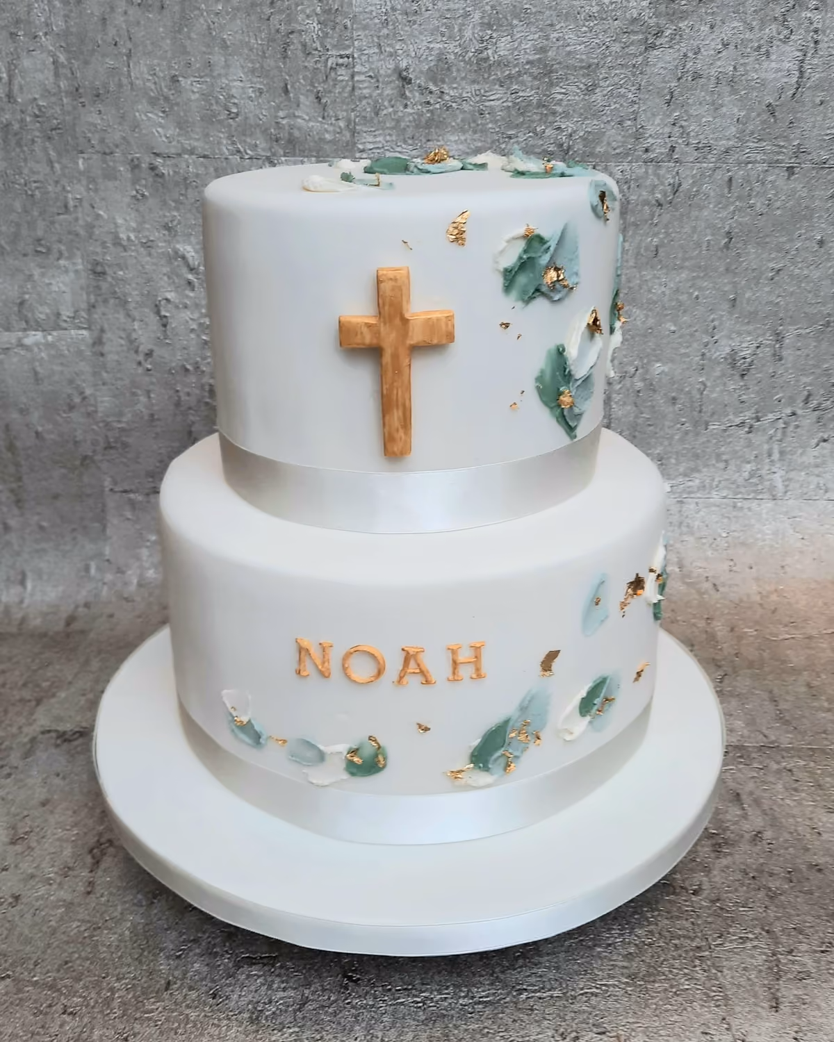 Two-tier white fondant christening cake featuring a golden cross on the top tier and the name 'NOAH' in gold letters on the bottom tier. Both tiers are decorated with abstract green, light blue, and white brushstroke details and gold leaf, with a white ribbon around the base of each.