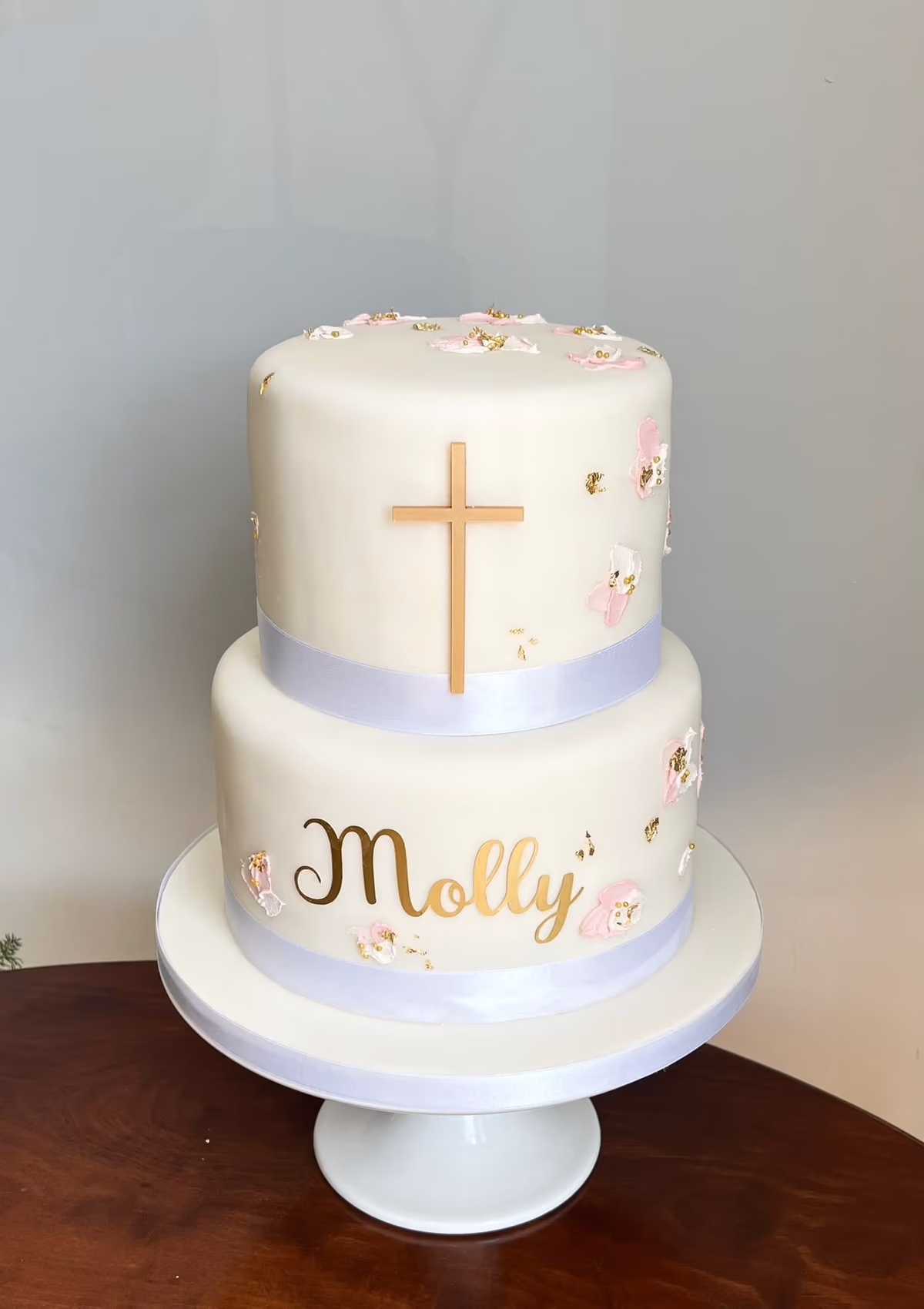 A two-tier white christening cake, decorated with a gold cross on the top tier and the name 'Molly' in gold script on the bottom tier. Both tiers feature delicate pink and white textured details with gold flecks and are encircled by a light lavender ribbon.