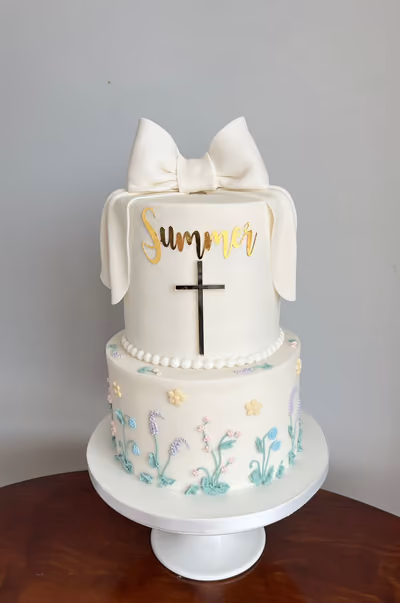 A two-tier white christening cake featuring a large white fondant bow, a gold 'Summer' name, and a gold cross on the top tier, with delicate pastel piped flowers decorating the bottom tier.