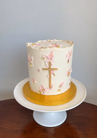 A single-tier white christening cake decorated with pink brushstroke details, gold leaf, edible pearls, and a prominent gold cross.