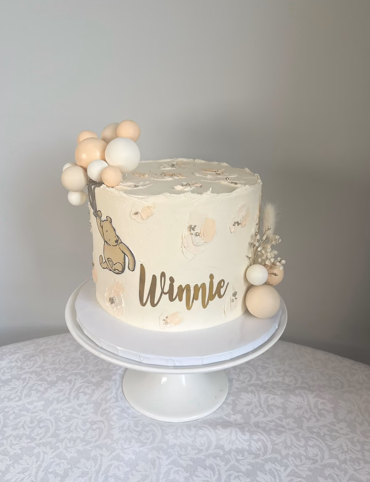 A cream buttercream christening cake for Winnie with a vintage Winnie the Pooh picture, gold 'Winnie' lettering, peach and white balloons and dried flowers.
