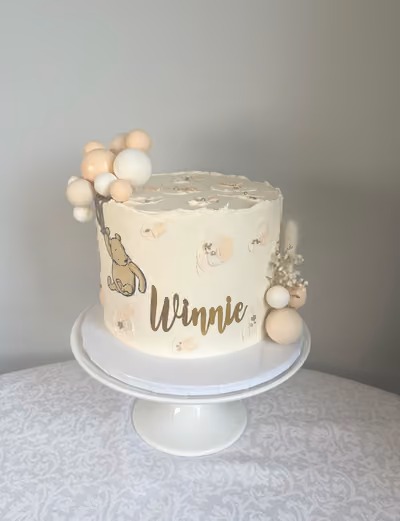 A cream buttercream christening cake for Winnie with a vintage Winnie the Pooh picture, gold 'Winnie' lettering, peach and white balloons and dried flowers.