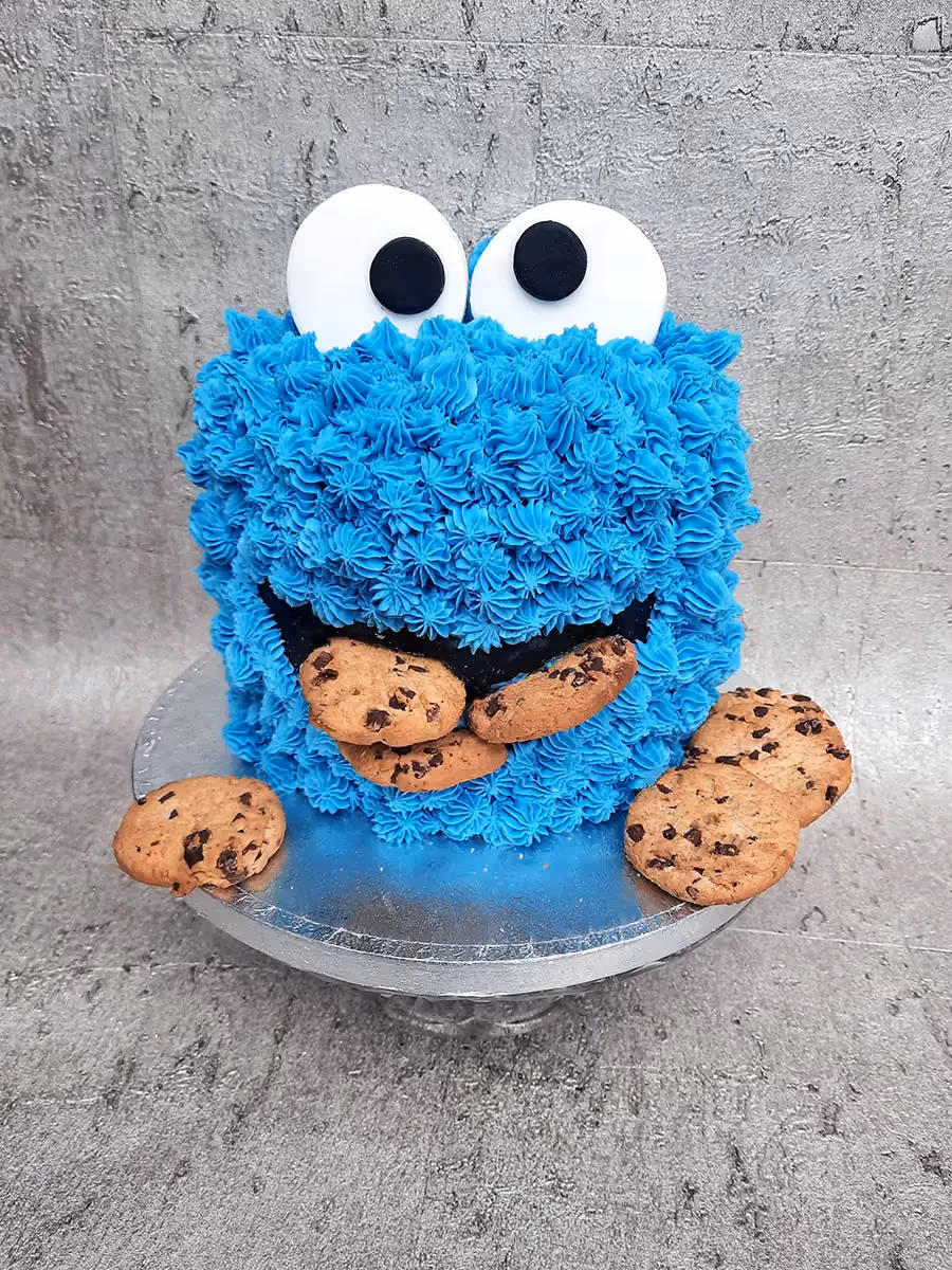 A vibrant blue Cookie Monster cake with large white and black eyes, decorated with several chocolate chip cookies around its mouth and on the silver cake stand.