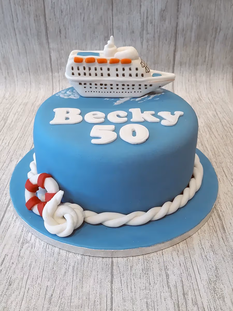 A round blue birthday cake decorated with a white cruise ship on top, the name 'Becky' and the number '50' in white fondant, and a white rope and red and white lifebuoy around the base.