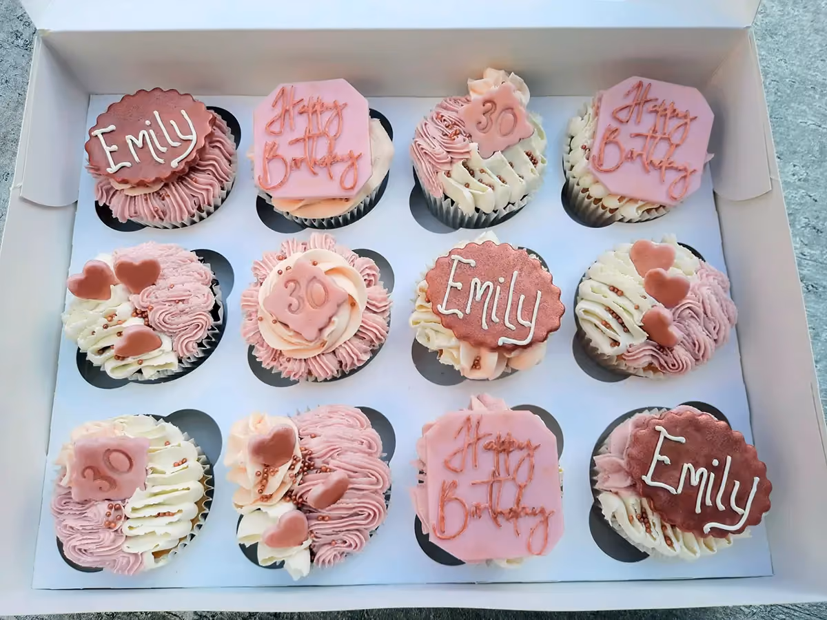 A dozen cupcakes in a white box, decorated with pink and white frosting, some featuring 'Emily', 'Happy Birthday', and '30' toppers, along with heart shapes and sprinkles, for a 30th birthday.