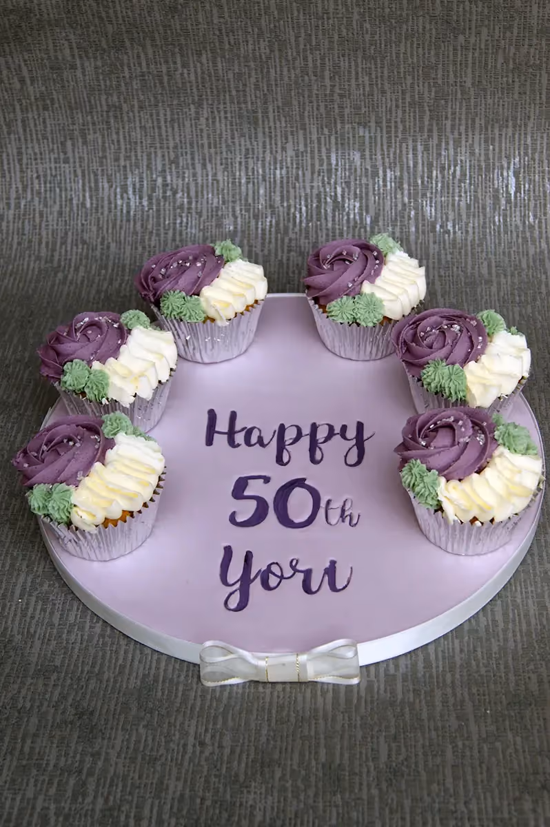 A round purple cake board with six cupcakes arranged in a circle. Each cupcake has purple and cream rose-style frosting with green leaves and silver sprinkles. The board is inscribed with 'Happy 50th Yori' in purple lettering and features a white ribbon bow at the bottom.
