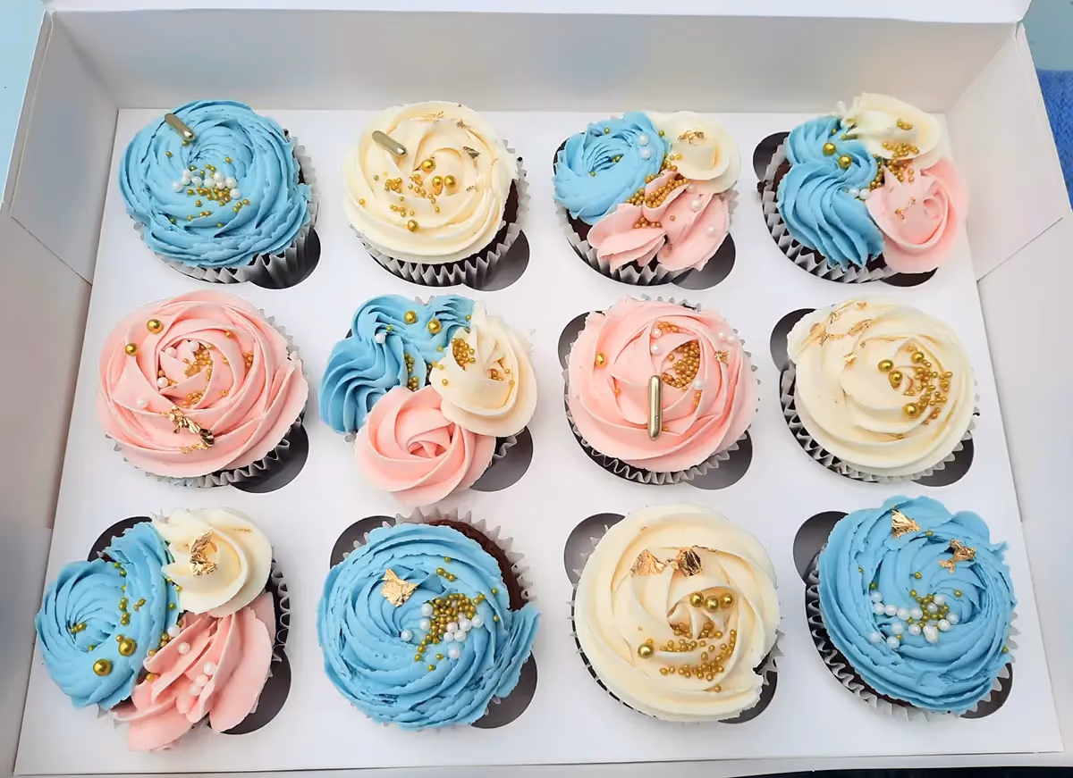 A dozen cupcakes in a white box, featuring various frosting designs in light blue, pink, and cream, all decorated with gold sprinkles and gold leaf accents.