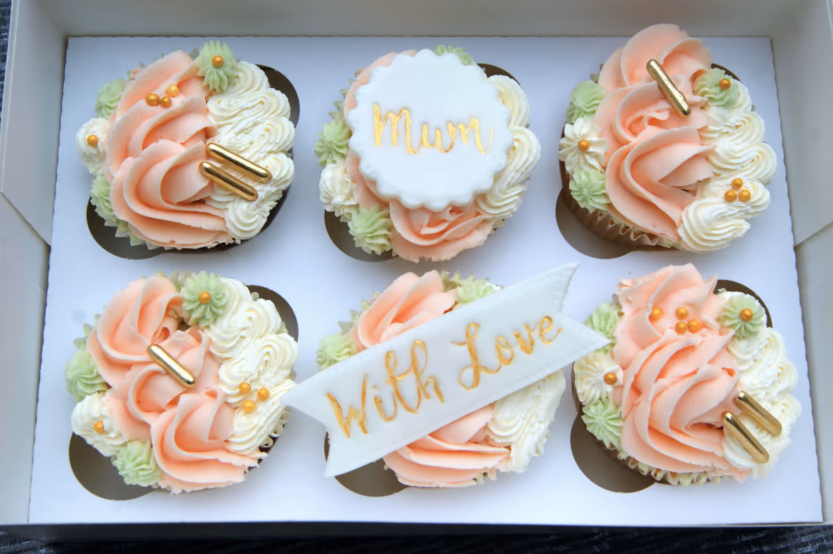 A box of six decorated cupcakes with peach, white, and light green frosting, gold sprinkles, and gold bar-shaped sprinkles. Two cupcakes feature white fondant toppers, one saying 'Mum' and another on a banner saying 'With Love' in gold script.