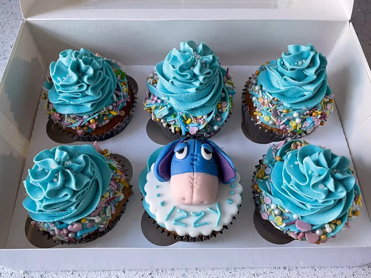 Six cupcakes presented in a white box; five have swirled teal frosting with colorful sprinkles, and one features an Eeyore character decoration with the name 'IZZY' written in light blue fondant.