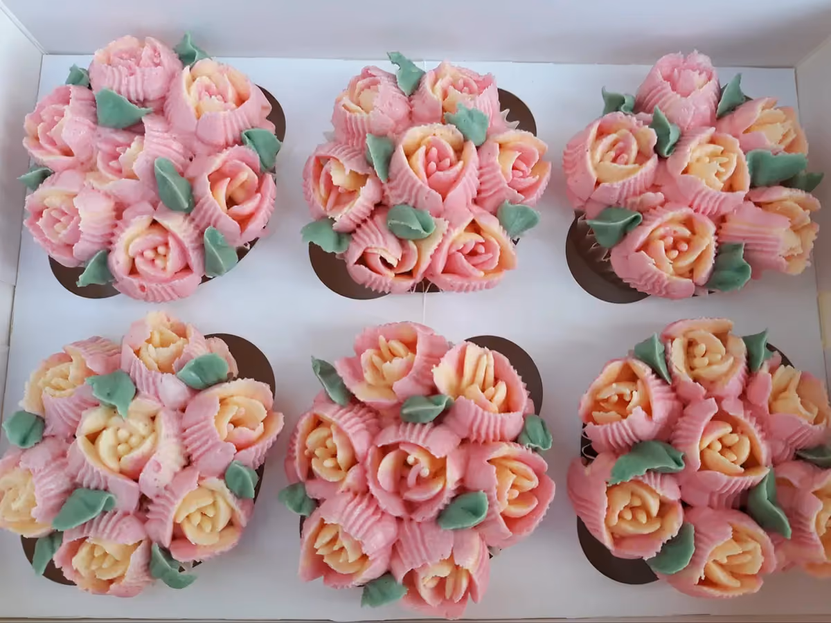Six cupcakes in a white box, each decorated with pink and yellow buttercream roses and green leaves, resembling small floral bouquets.