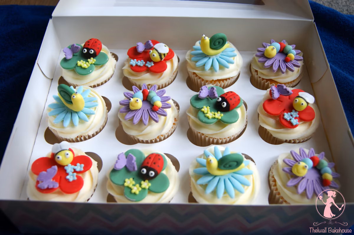 A white box containing 12 garden-themed cupcakes, each decorated with a different colorful fondant bug or creature such as ladybugs, bees, snails, and caterpillars, all on various colored flowers.