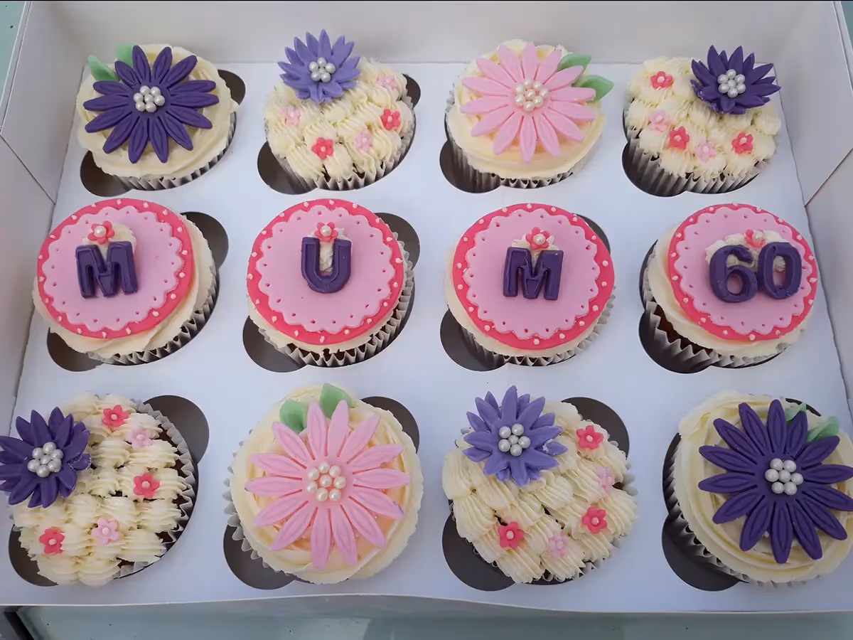 Twelve cupcakes in a white box, featuring pink and purple decorations. Some cupcakes spell 'MUM', others display '60', and the remaining are adorned with purple or pink fondant flowers on white or cream frosting.