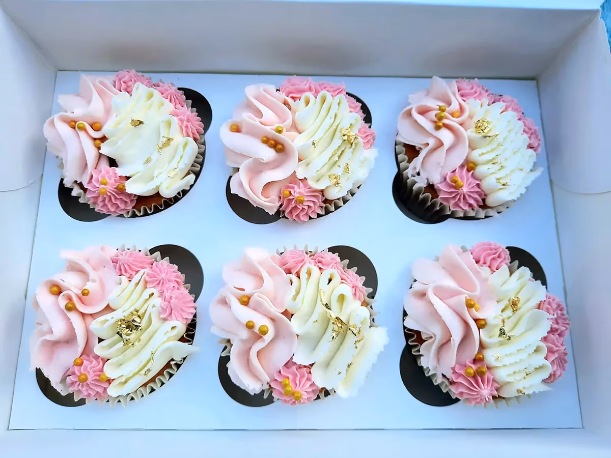 Six cupcakes in a white box, each decorated with swirled pink and white buttercream frosting, gold leaf, gold sprinkles, and small piped pink flowers.