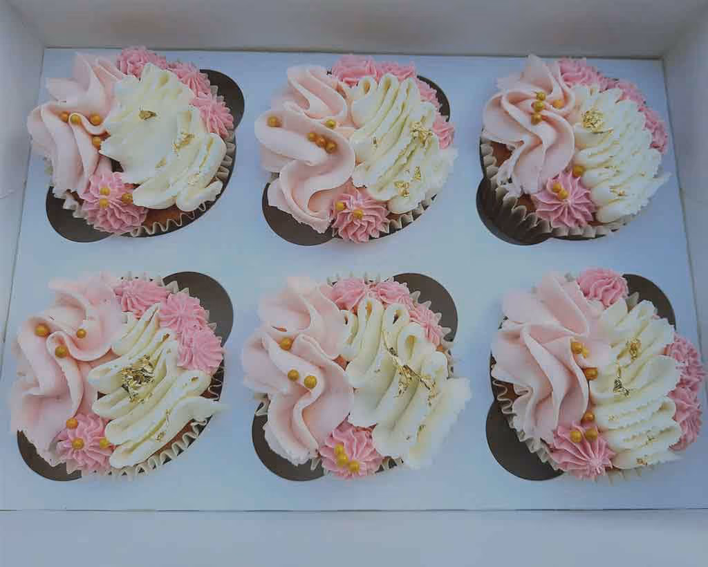 Six cupcakes arranged in a white box, each decorated with swirled pink and white frosting, small pink piped flowers, gold leaf flakes, and small gold spherical sprinkles.