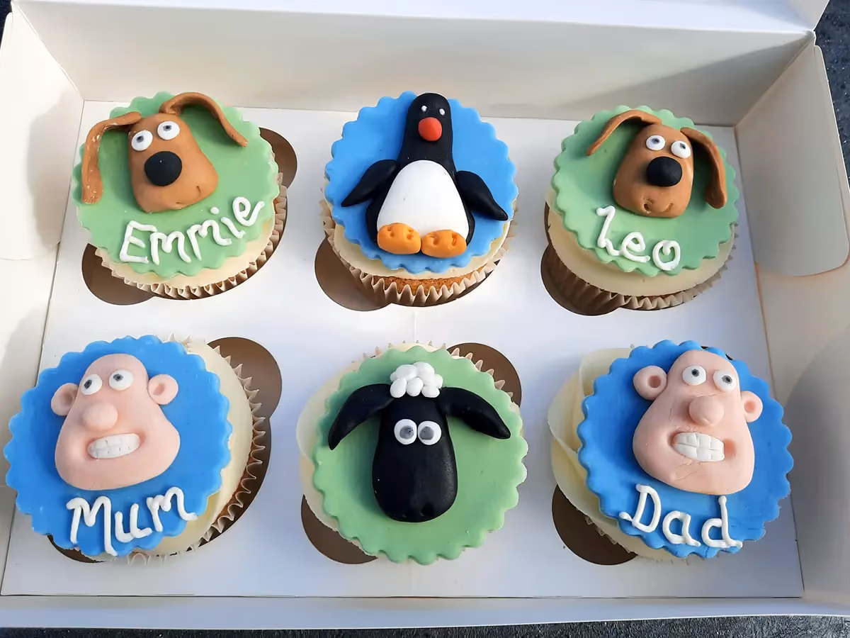 Six cupcakes in a white box, decorated with fondant characters from Wallace and Gromit and Shaun the Sheep. The top row features two brown dog heads named 'Emmie' and 'Leo', and a penguin. The bottom row features two pink human faces named 'Mum' and 'Dad', and a black sheep.
