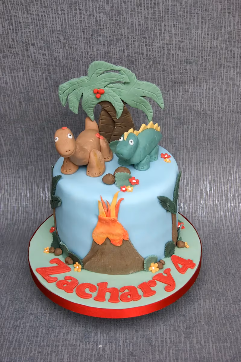 Single-tier light blue dinosaur-themed birthday cake for Zachary's 4th birthday, featuring two fondant dinosaurs, a palm tree, and a volcano with lava, all on a light green base with red lettering.
