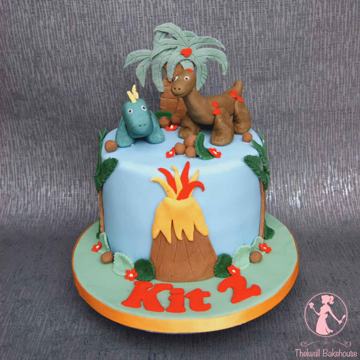 A light blue round birthday cake decorated with two fondant dinosaurs, a volcano, palm trees, and red flowers, with 'Kit 2' written in red on the green base.