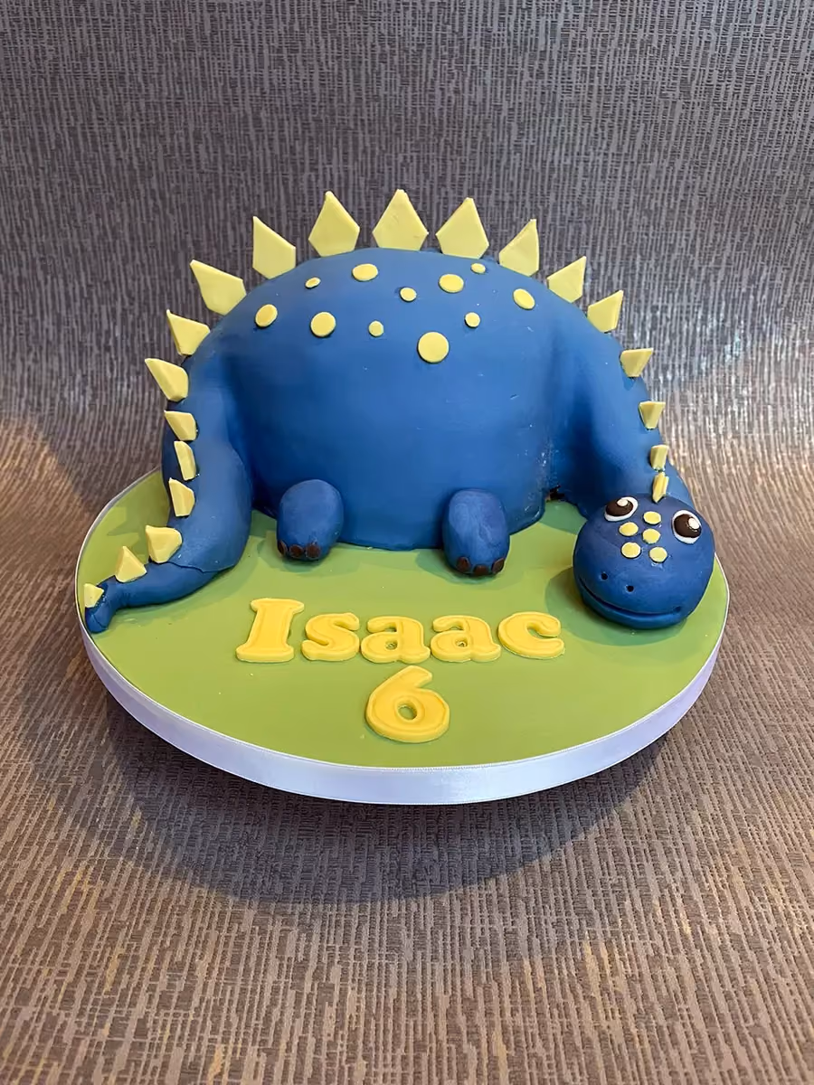A blue dinosaur-shaped birthday cake with yellow spikes and polka dots, featuring the name 'Isaac' and the number '6' in yellow on a green base.
