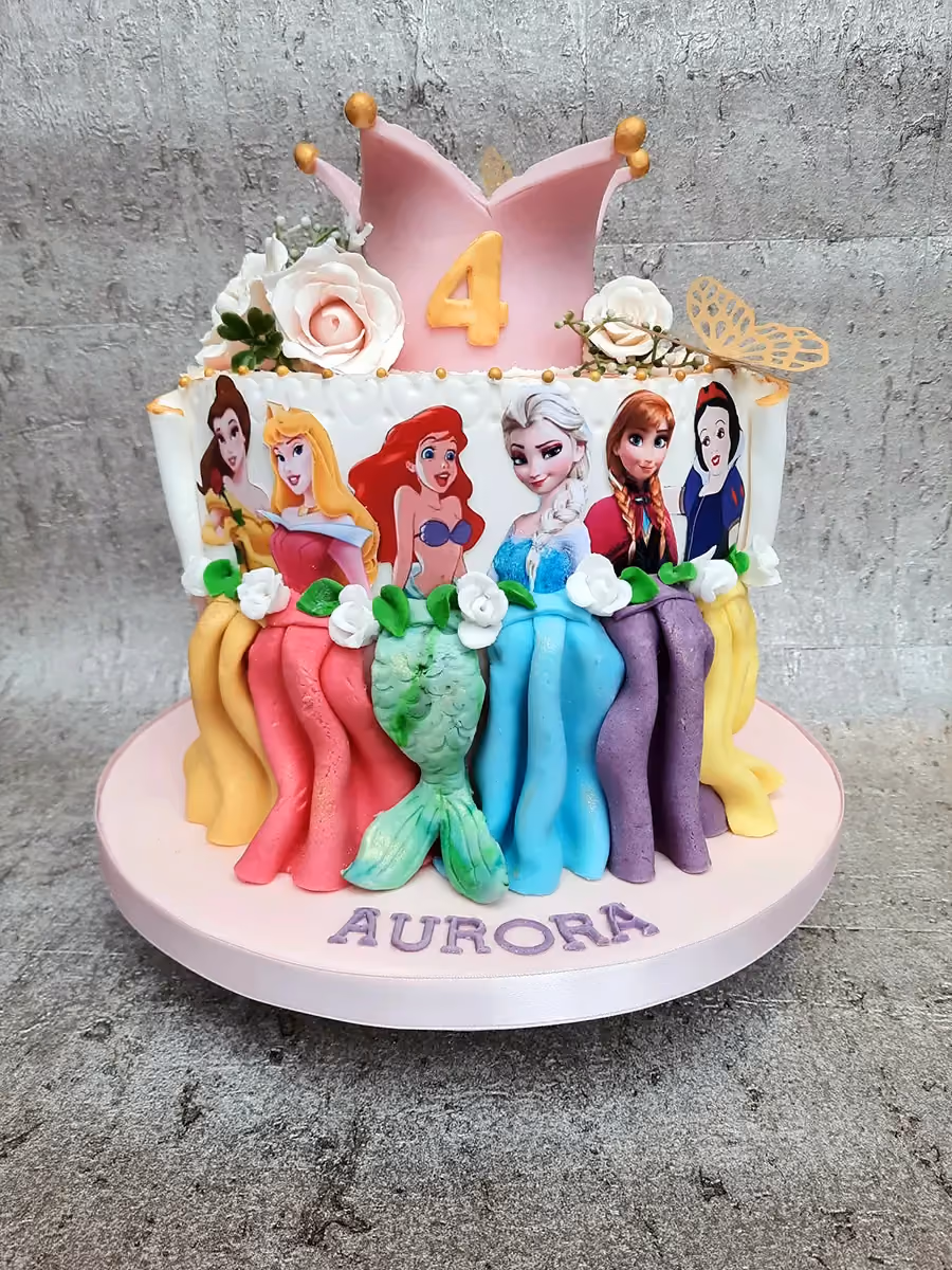 A vibrant Disney Princess birthday cake featuring images of Belle, Aurora, Ariel, Elsa, Anna, and Snow White, topped with a pink crown displaying the number 4, sugar flowers, a golden butterfly, and the name 'Aurora' written on the base.