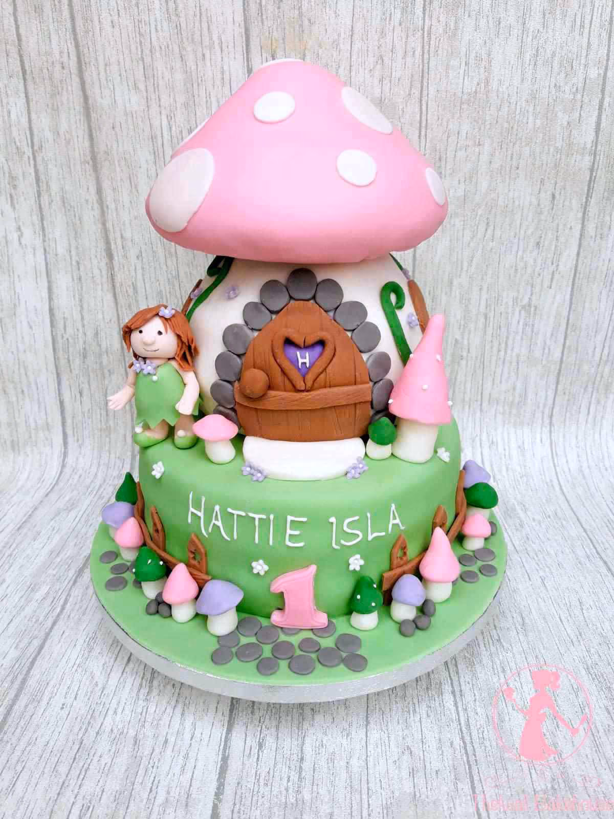 A two-tier fairy mushroom house themed first birthday cake. The top tier is a pink and white spotted mushroom cap over a white house with a brown door featuring a heart-shaped window and an 'H' initial. The bottom green tier is decorated with a fairy figurine, various small pink, purple, and green mushrooms, and the white text 'HATTIE ISLA' and a pink number '1'.