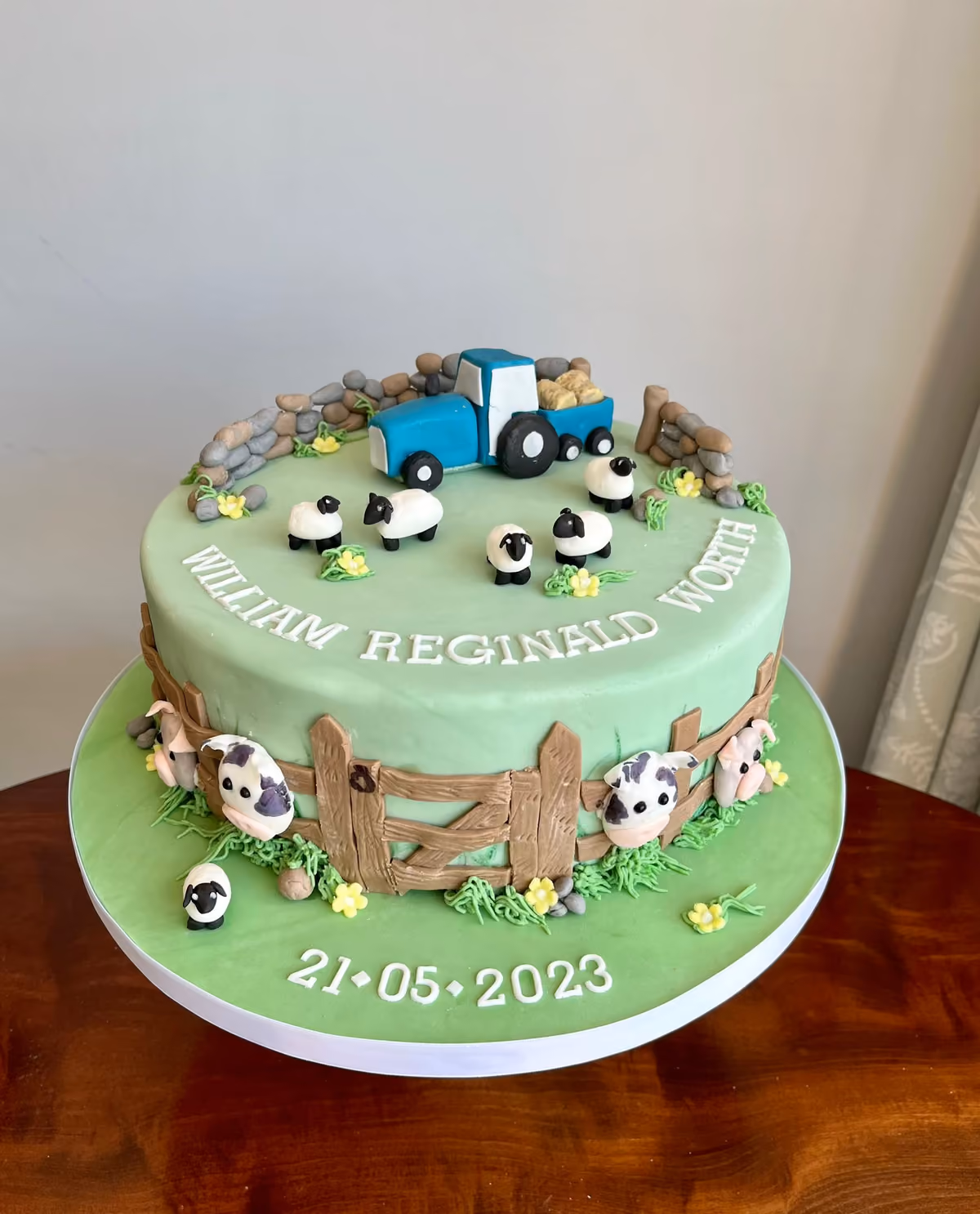 A two-tier farm-themed cake with light green fondant, featuring a blue tractor with hay, sheep, and stone walls on the top tier. The bottom tier is decorated with a brown wooden fence, cows, a pig, and a lamb. The cake displays the name 'WILLIAM REGINALD WORTH' and the date '21-05-2023'.