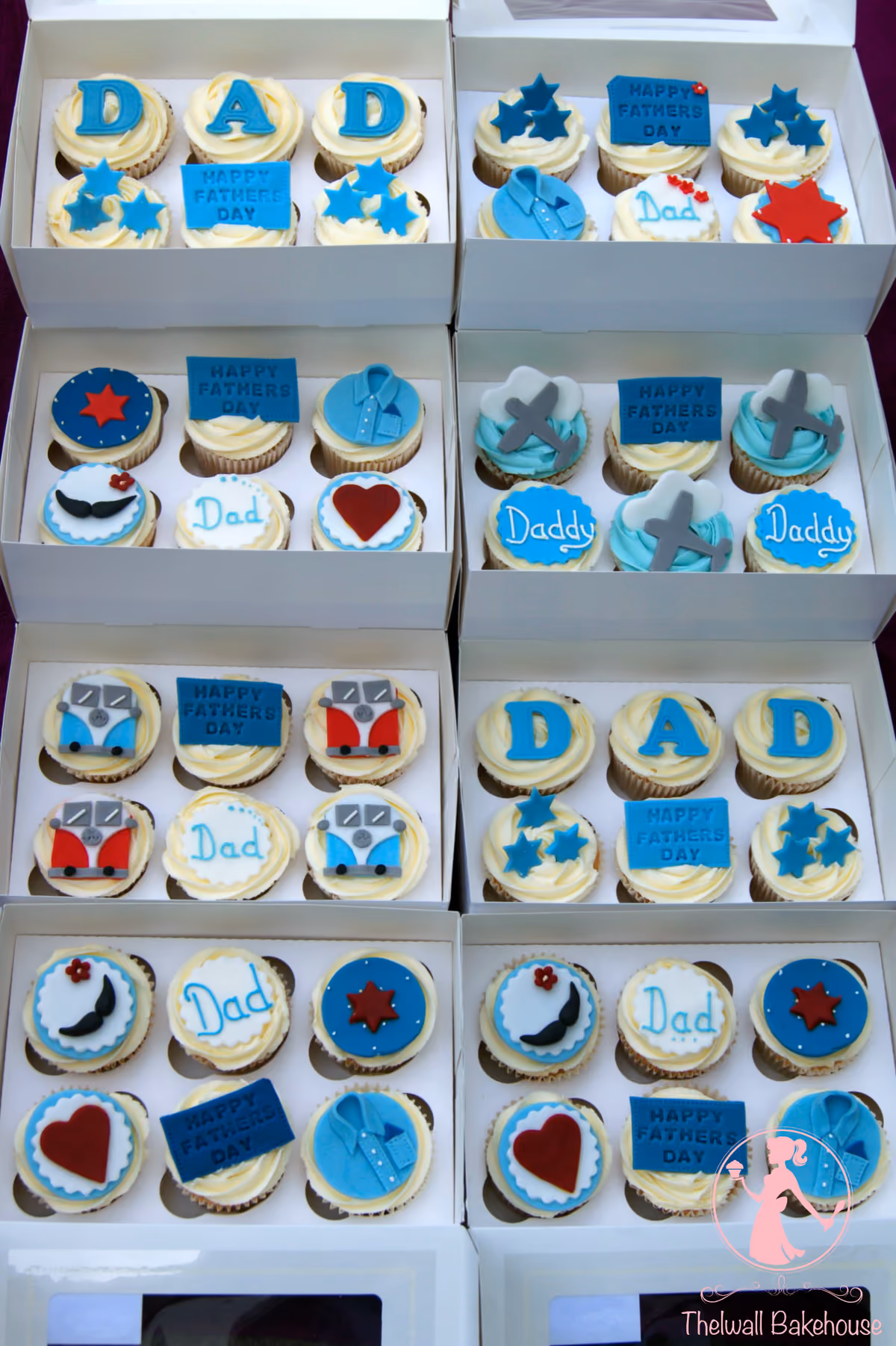 An overhead view of multiple white boxes filled with Father's Day themed cupcakes. The cupcakes feature various designs including blue 'D-A-D' letters, 'Happy Fathers Day' plaques, blue shirts, stars, red hearts, mustaches, airplanes, and colorful camper vans, primarily in blue, red, and white frosting.
