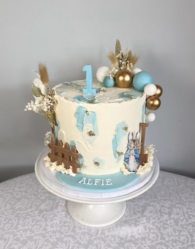 A Peter Rabbit first birthday cake for Alfie with pale blue brushstrokes, a wooden fence, a blue '1' topper, dried flowers and gold, white and blue spheres.