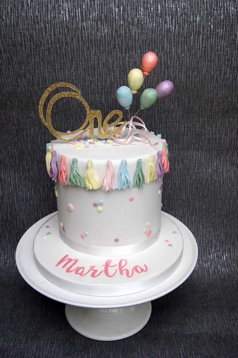 A single-tier white fondant first birthday cake decorated with a glitter gold 'One' topper, pastel balloons, pastel tassels, colorful sprinkles, and the name 'Martha' written in pink on the cake board.