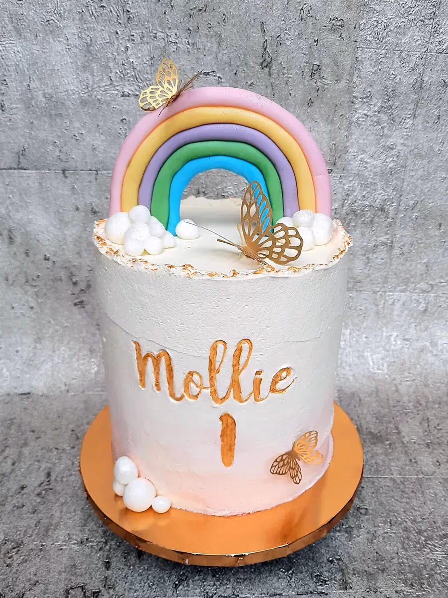 A single-tier white and pink ombre birthday cake for Mollie's first birthday, decorated with a pastel rainbow arch, white clouds, and gold butterfly accents.