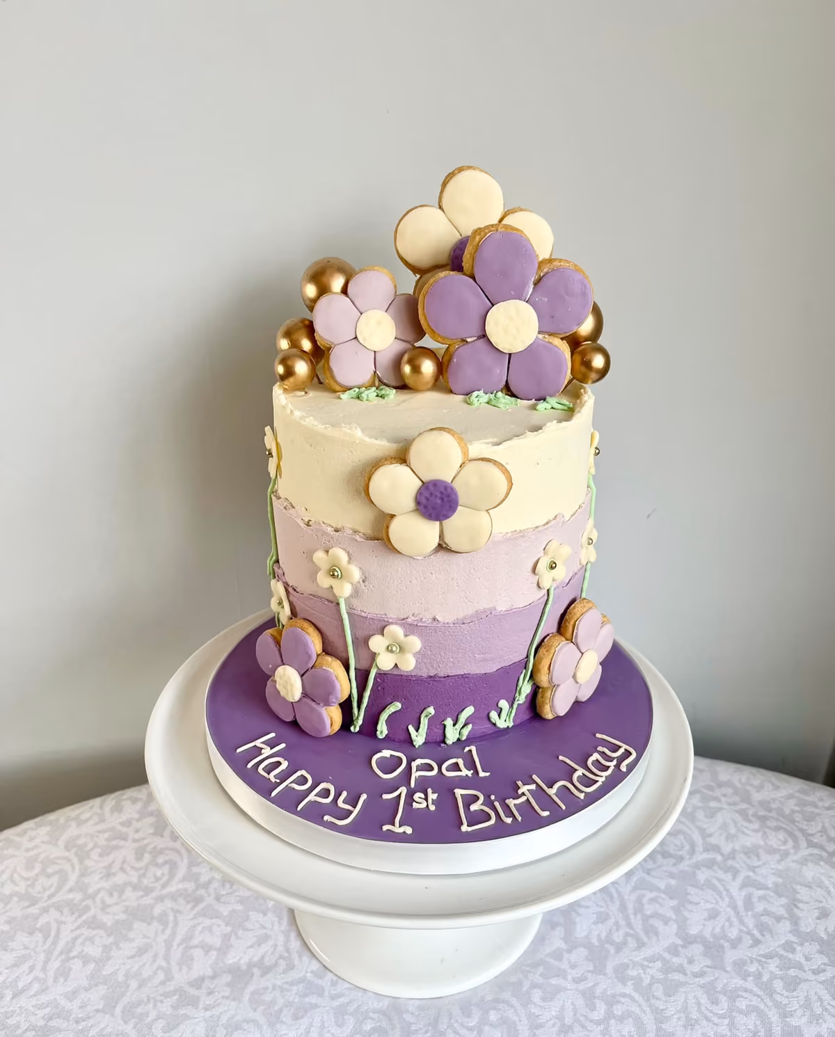 A purple ombre buttercream first birthday cake for Opal decorated with flower-shaped biscuits in lilac, purple and cream, piped stems and gold spheres.