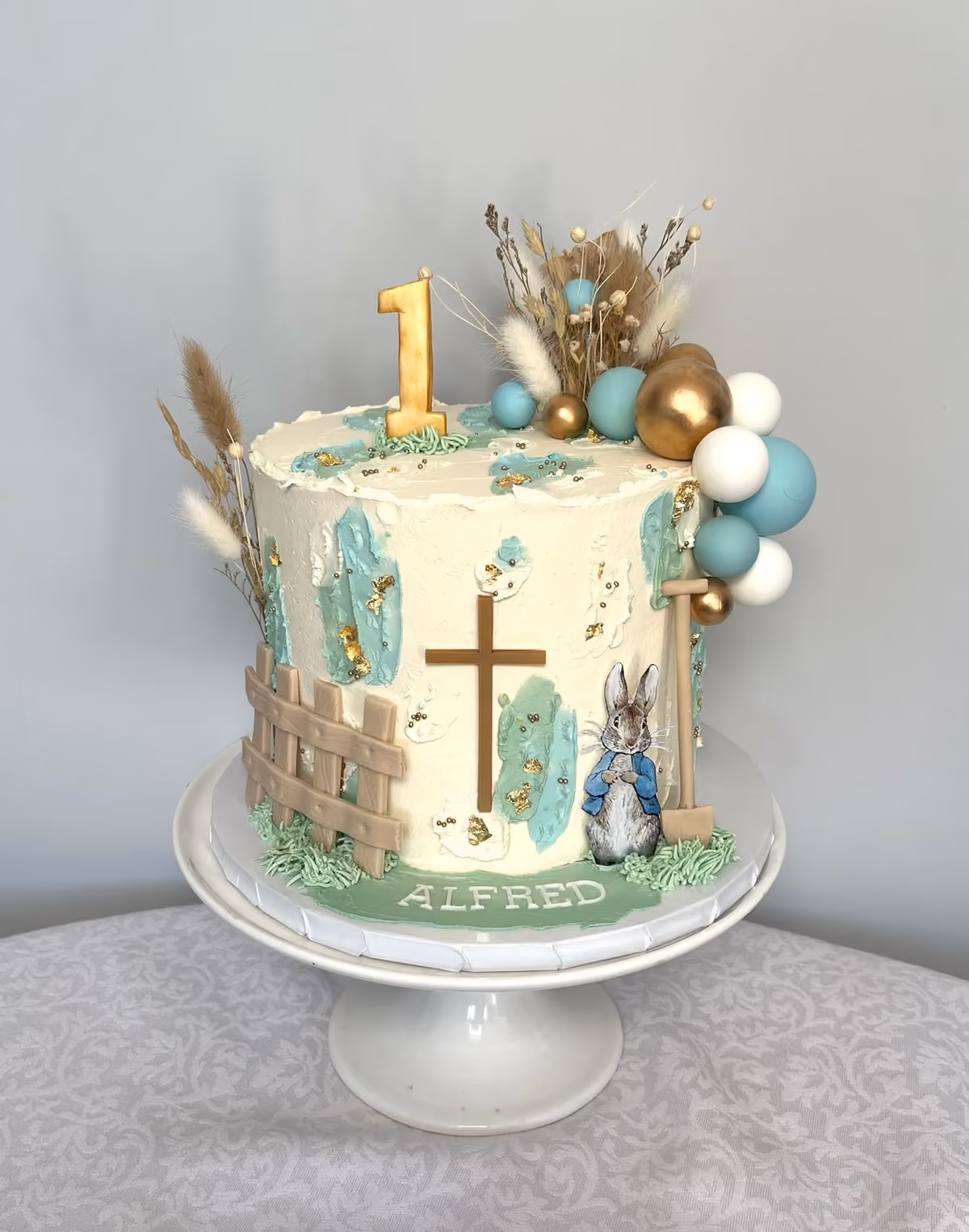 A Peter Rabbit themed first birthday cake for Alfred, decorated with a gold number one candle, a brown cross, a wooden fence, a Peter Rabbit figure, dried floral arrangements, and teal, gold, and white spherical decorations, all on a white buttercream cake with teal brush strokes and gold leaf.
