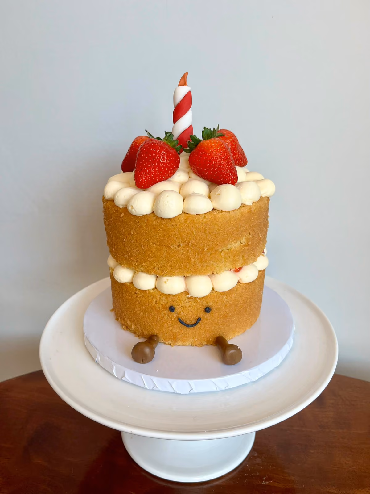 A cute smiling first birthday cake made to look like a sponge cake with cream and strawberries, little legs and a striped candle.