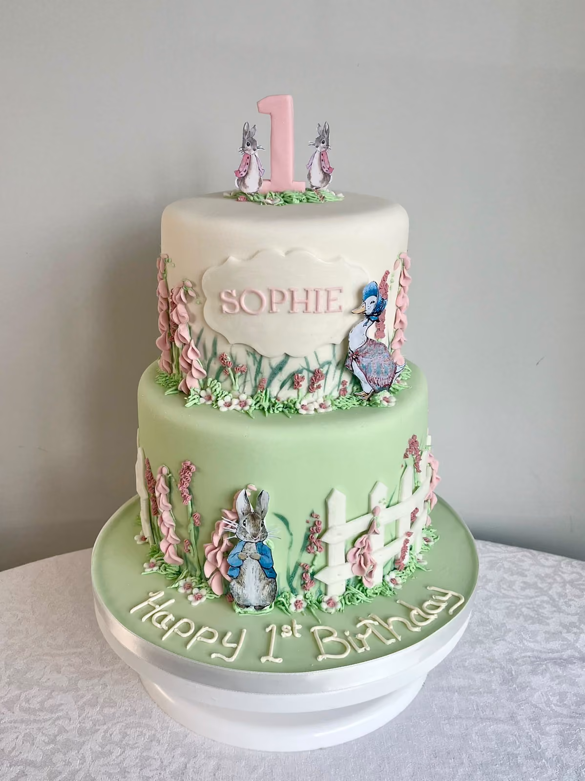A two-tier Peter Rabbit first birthday cake for Sophie in white and sage green, with Peter Rabbit and Jemima Puddle-Duck, pink foxgloves and a white fence.