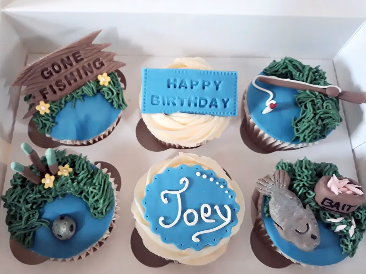 Six fishing-themed birthday cupcakes are displayed in a white box. Decorations include a 'GONE FISHING' sign, a blue 'HAPPY BIRTHDAY' plaque, a fishing rod, cattails with a lure, a blue plaque with 'Joey' written in white, and a grey fish next to a 'BAIT' pot with pink worms. Most cupcakes feature blue water and green grass elements.