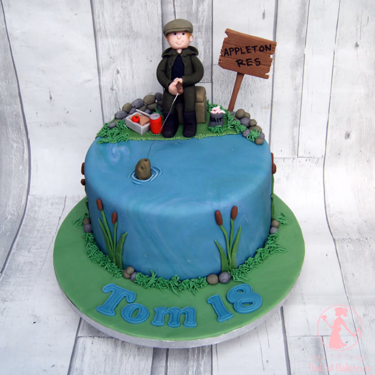 A round, single-tier birthday cake decorated as a fishing scene, featuring a fondant figure of an angler in a green jacket and cap sitting by a blue pond with a fishing rod and bobber. The cake includes details like a 'APPLETON RES' sign, fishing bait, cattails, and 'Tom 18' written in blue fondant letters on the green cake board.