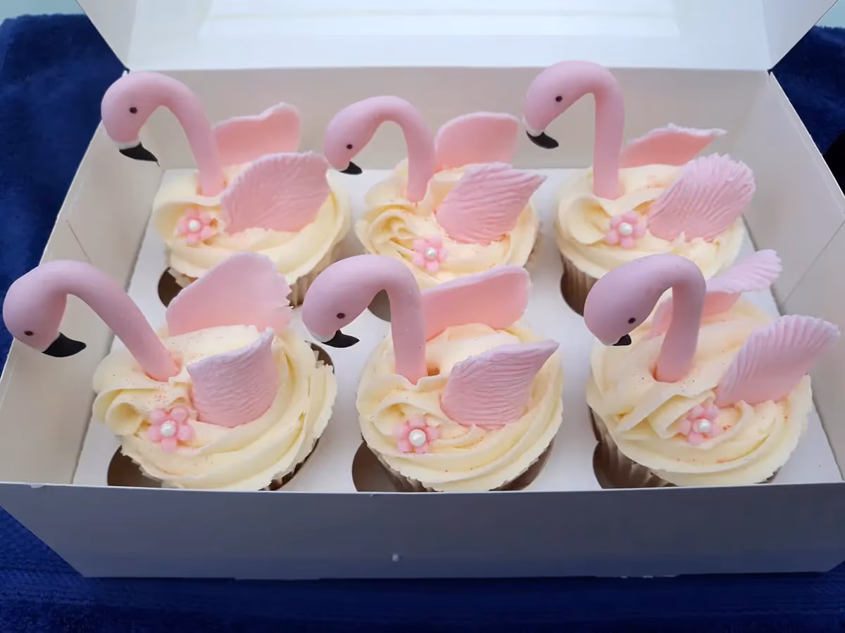 Six cupcakes decorated with pink flamingo heads, wings, and small pink flowers, presented in a white box.