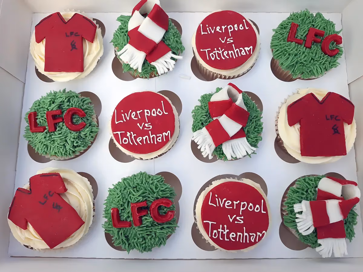 A dozen football-themed cupcakes arranged in a white box, featuring designs like red Liverpool FC shirts, red and white scarves, red circles with 'Liverpool vs Tottenham' text, and green grass frosting with red 'LFC' letters.