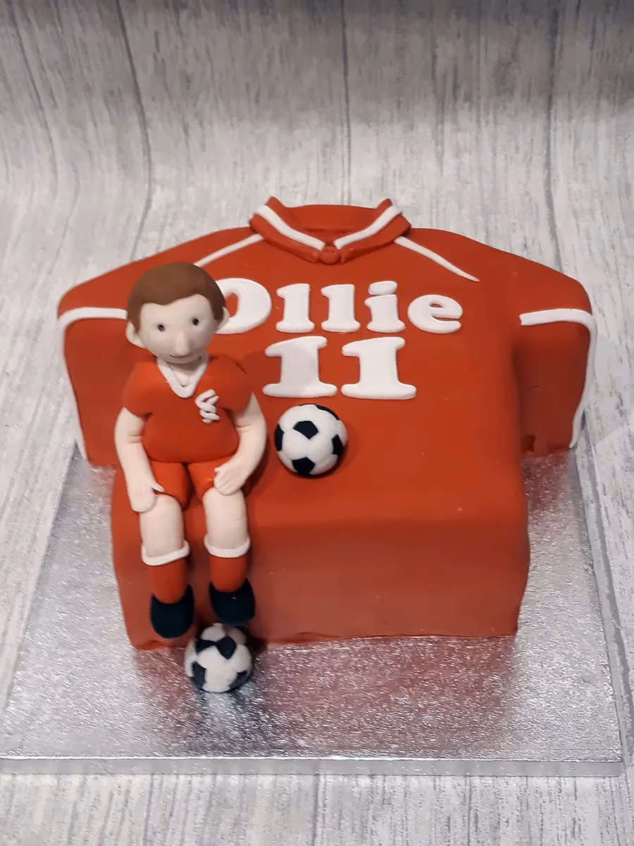 A red football shirt shaped cake with 'Ollie 11' written in white fondant, featuring a sugar paste figure of a boy in a red football kit sitting on the cake, with two small footballs.
